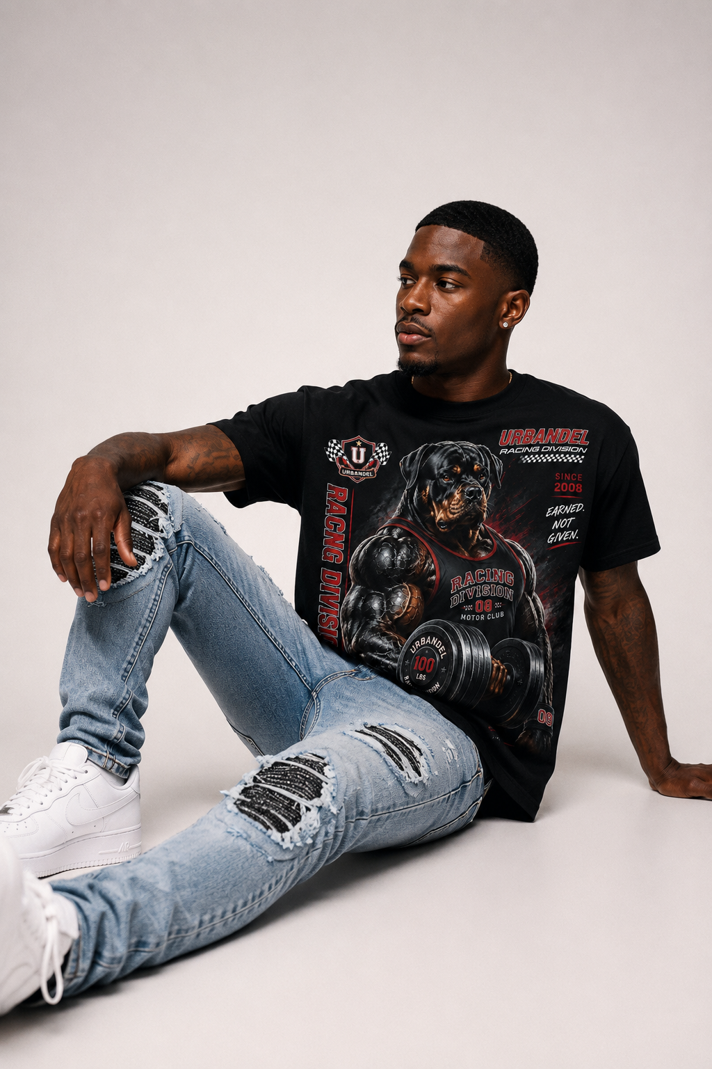 A man sitting on a light gray floor wearing a black graphic t-shirt with a muscular dog design, ripped light-wash blue jeans, and white sneakers.