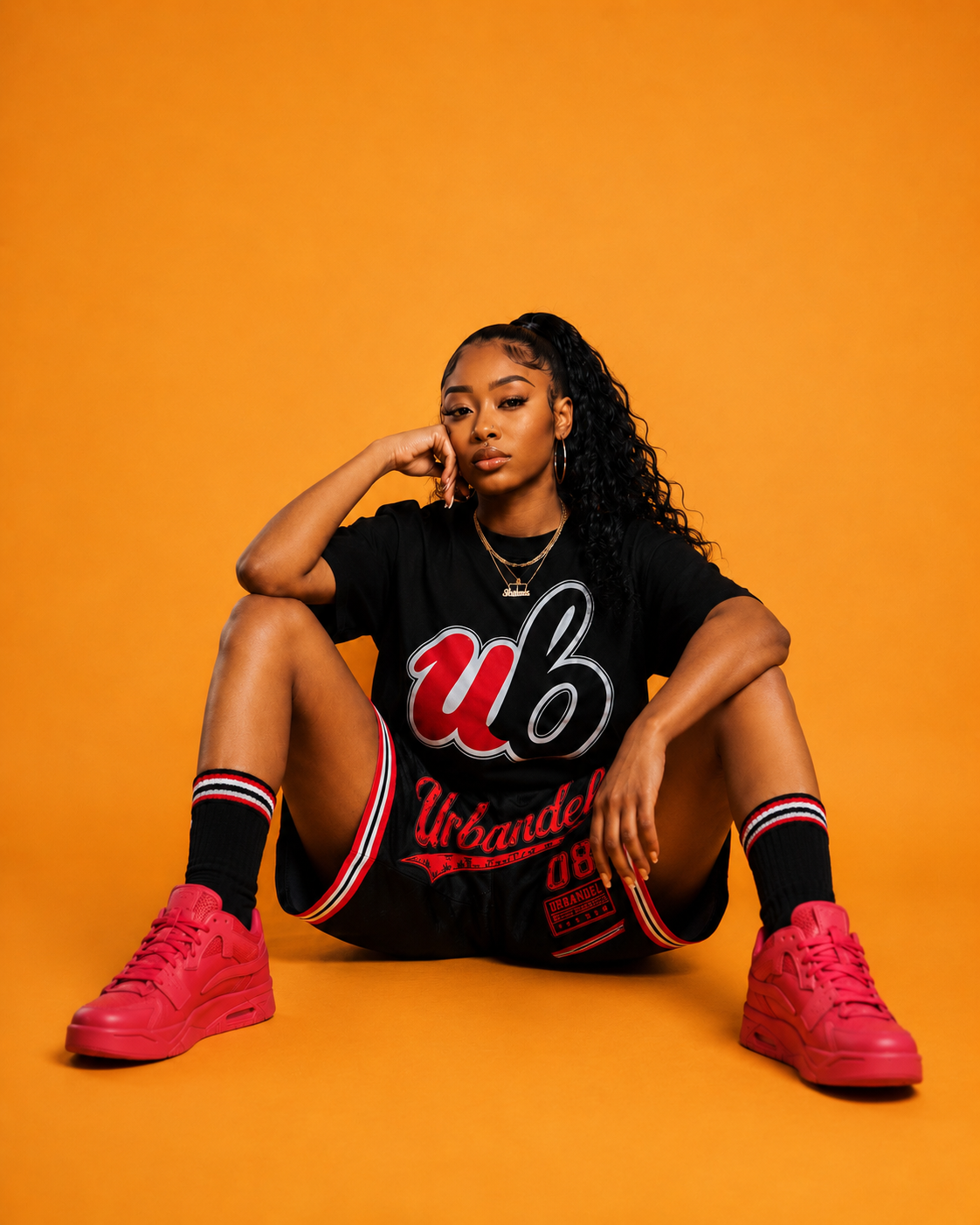 A woman sits on a solid orange studio floor wearing a black short-sleeve t-shirt with a red and white 'ub' graphic, matching black shorts, black socks with red and white stripes, and bright pink sneakers.